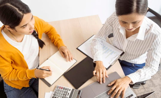 Two women working at a desk with a laptop and notebook, photo.
