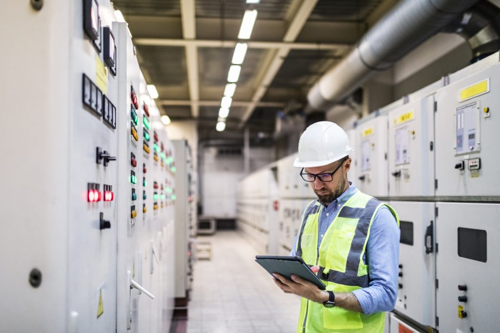 Engineer in safety gear using tablet in control room with lit panels.
