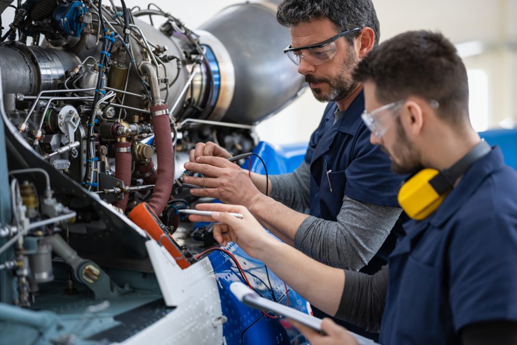 Two technicians work on aircraft engine wiring, wearing blue uniforms and goggles.