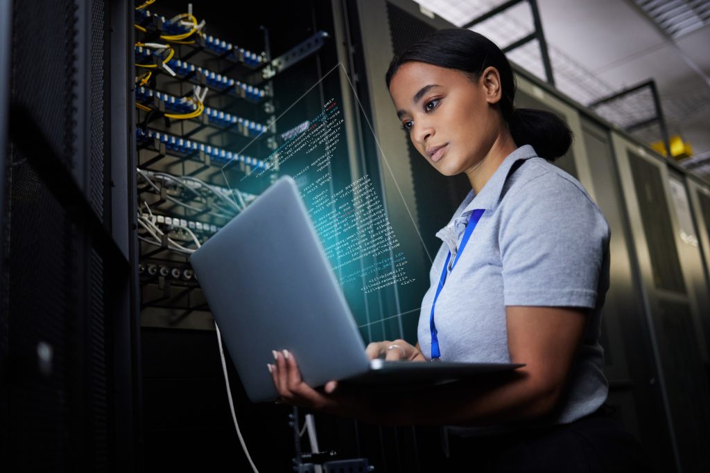 Woman working on laptop in server room, wearing gray shirt, digital illustration.